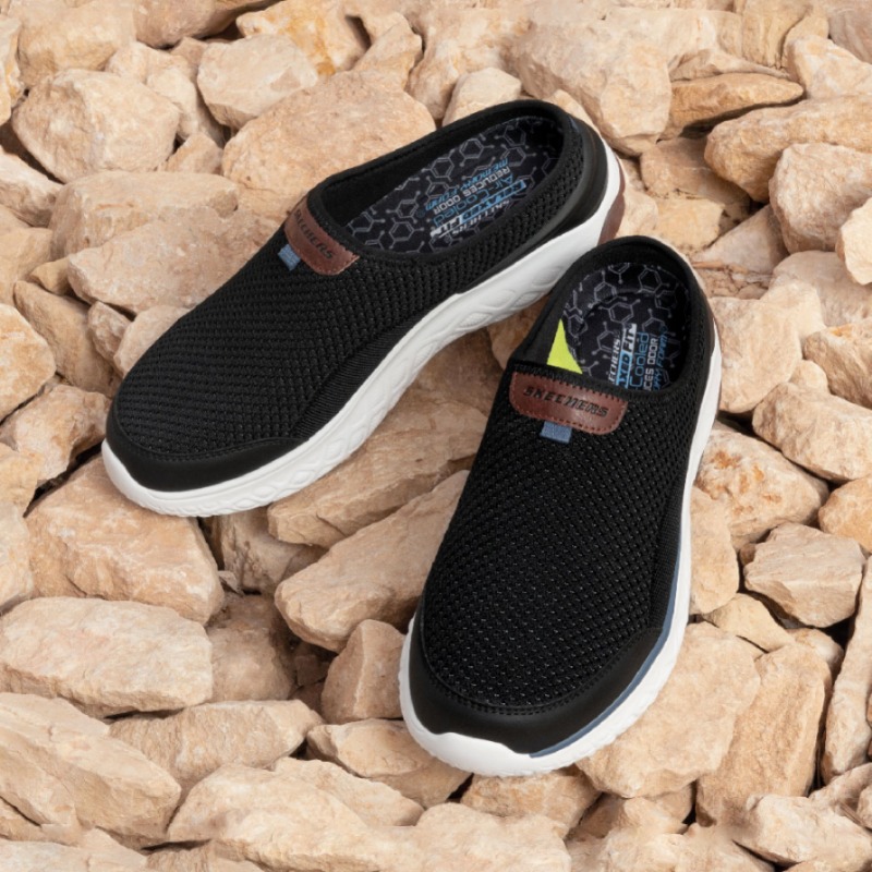A pair of black Skechers slip-on shoes with white soles resting on a bed of rocks.