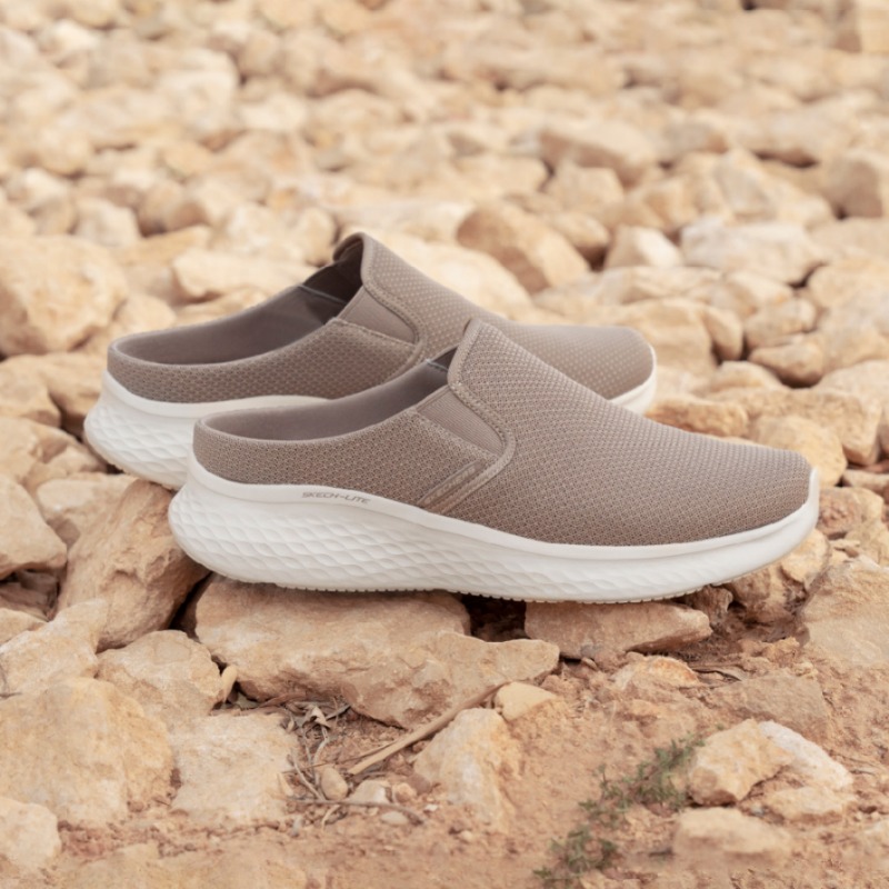 A pair of taupe Skechers slip-on mules with white soles resting on rocks.