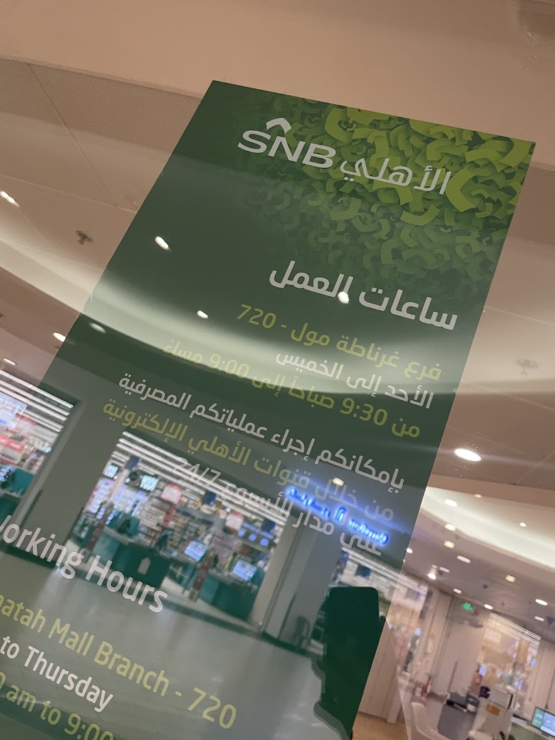 A sign displays SNB AlAhli bank's working hours and services, with a blurred view of the bank interior.