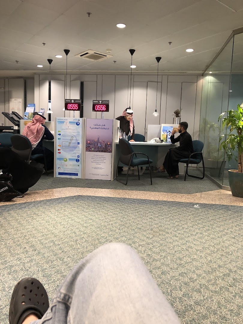 People at counters in a bank or service center with digital displays showing numbers.