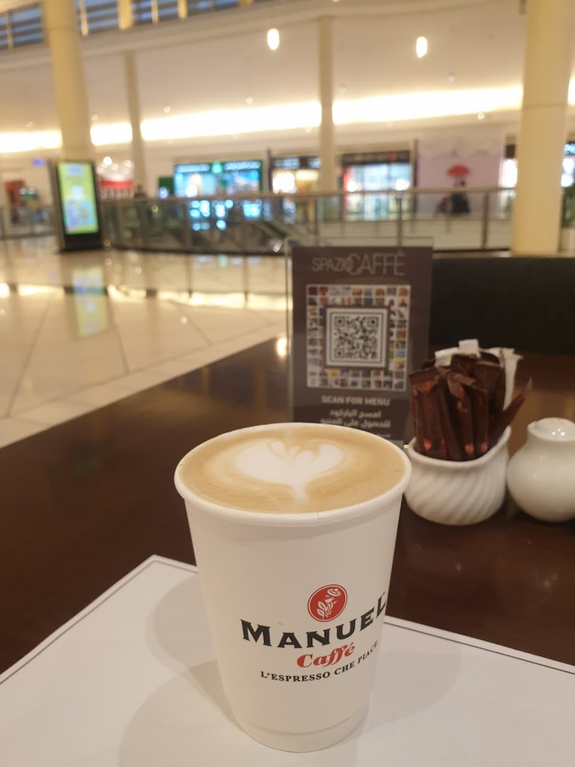 A cup of latte with heart art sits on a table in a cafe, with a mall interior in the background.