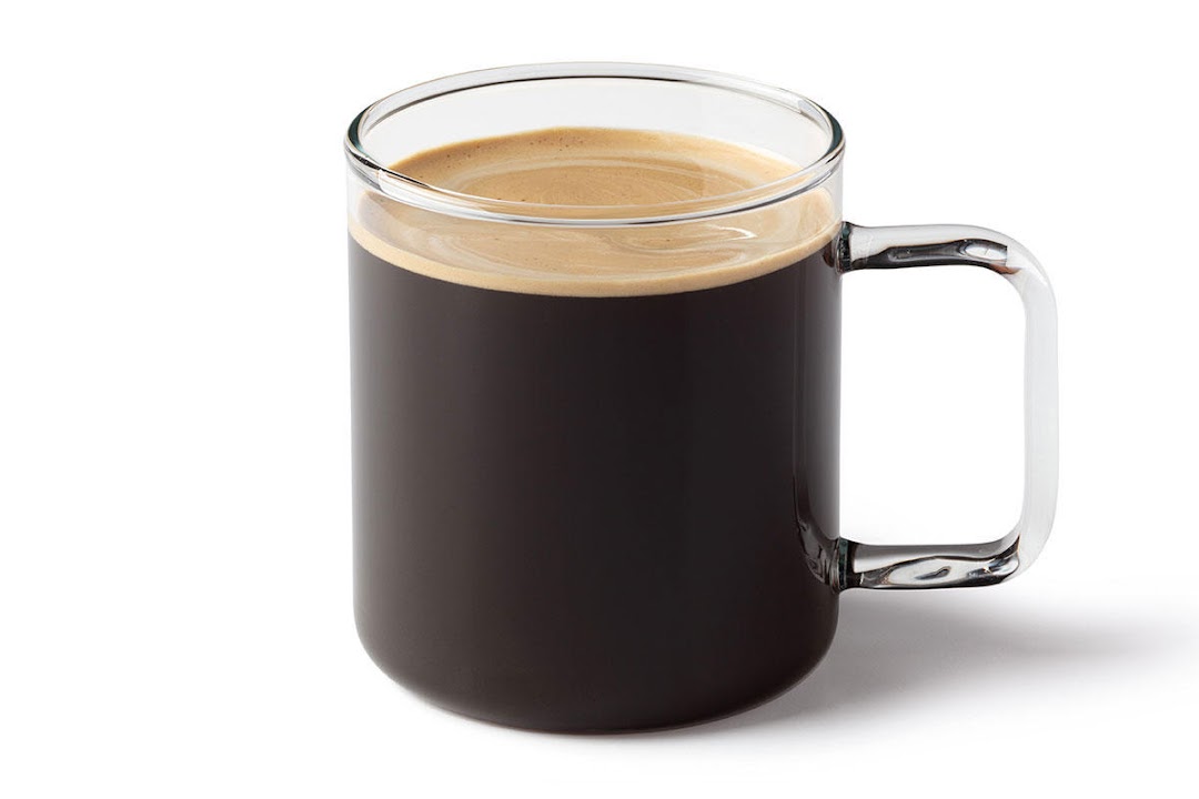 A clear glass mug filled with dark coffee and a layer of foam.