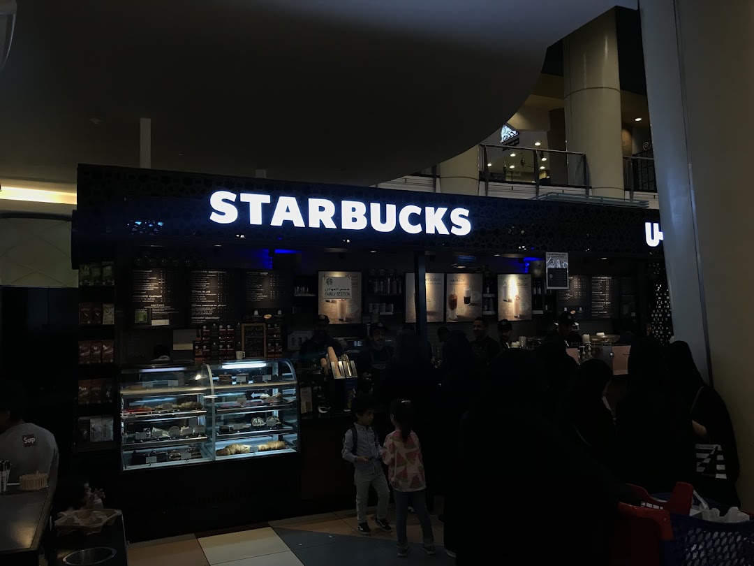 A Starbucks coffee shop with a display of pastries and a menu board, with customers queuing.