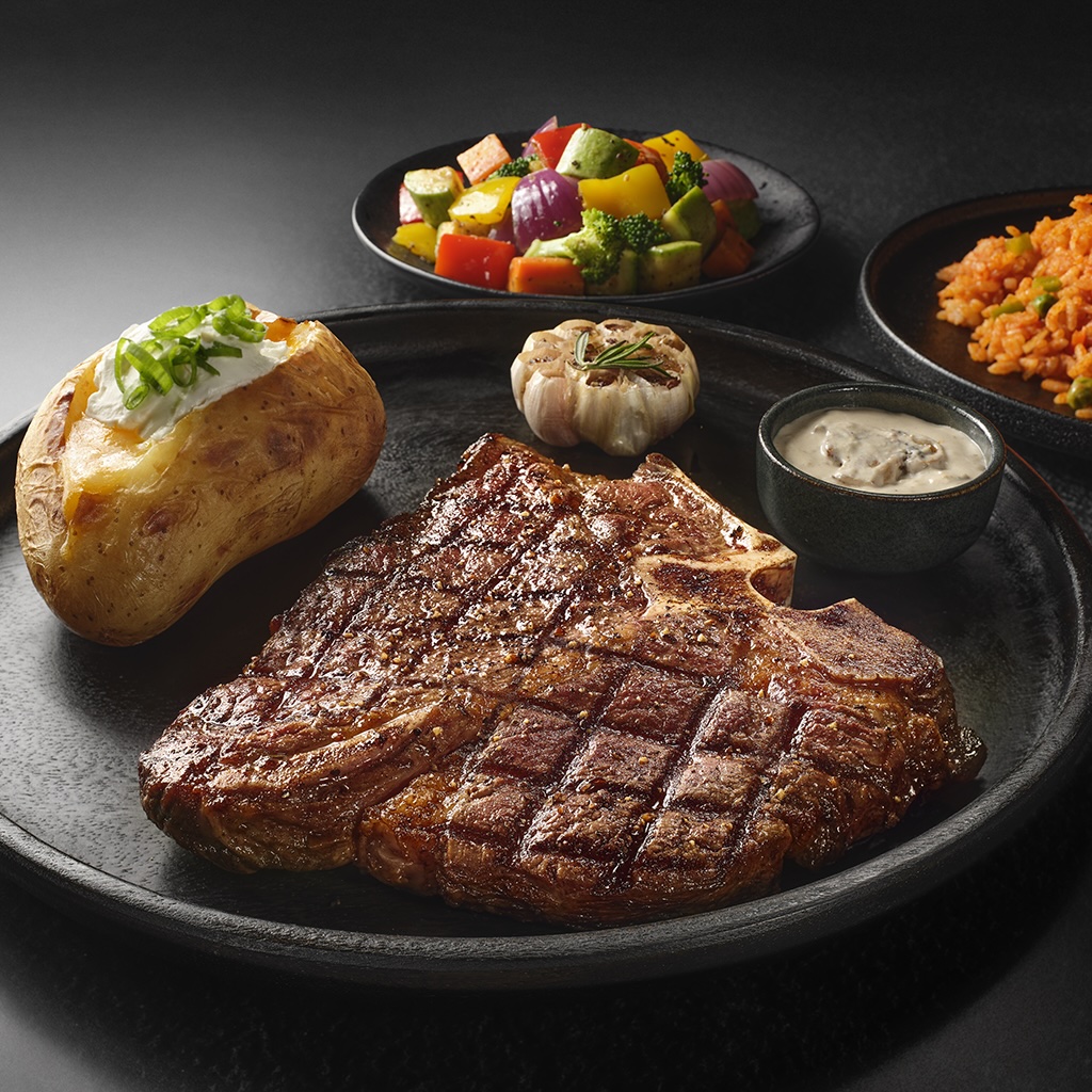 A perfectly grilled T-bone steak served with a baked potato, roasted garlic, and mixed vegetables.