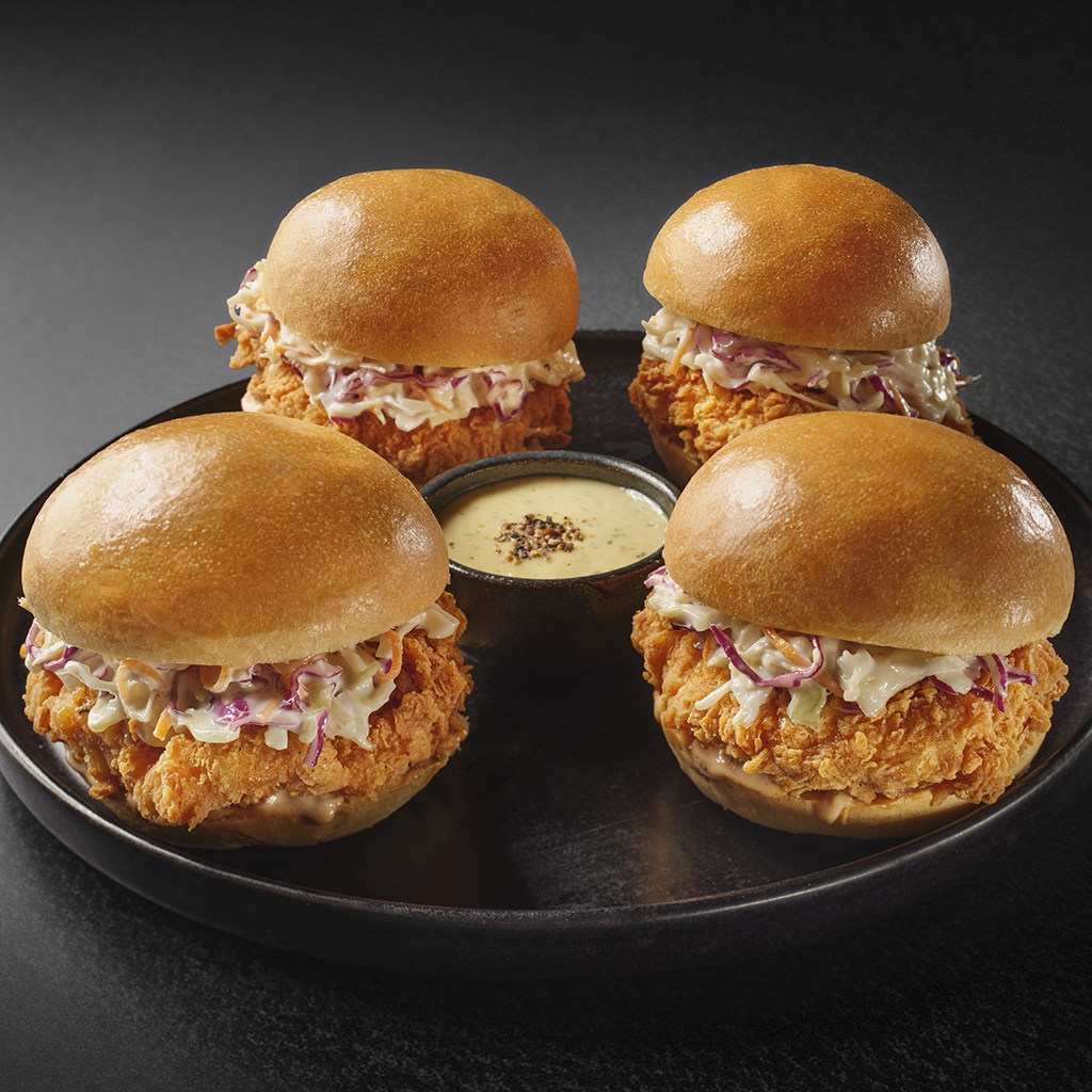Four crispy chicken sliders with coleslaw and a dipping sauce on a black plate.