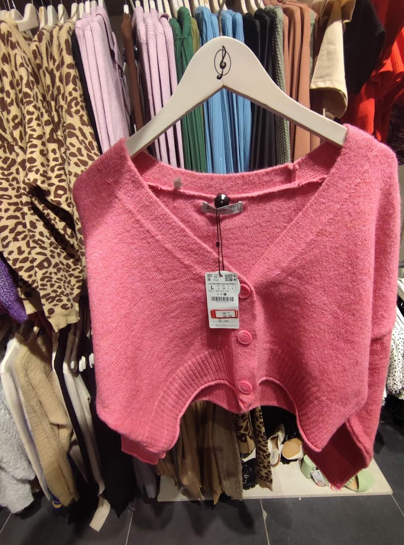 A pink knitted cardigan with buttons is displayed on a hanger, surrounded by other clothing items.