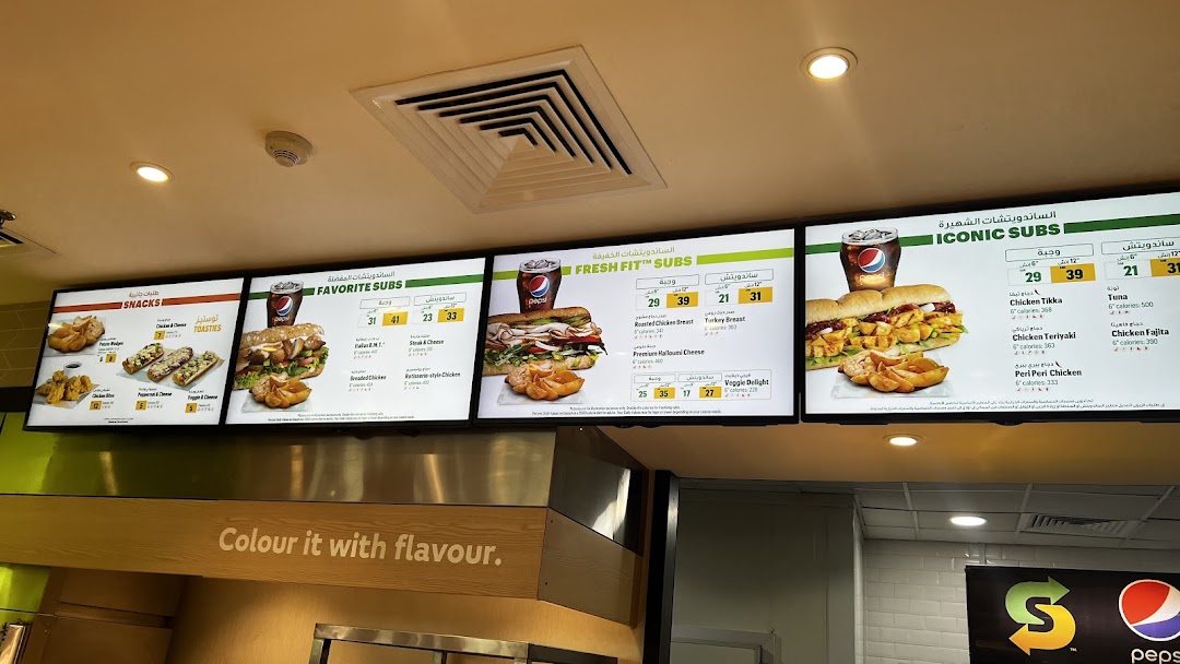 A digital menu board displaying various Subway subs, snacks, and drinks with prices.