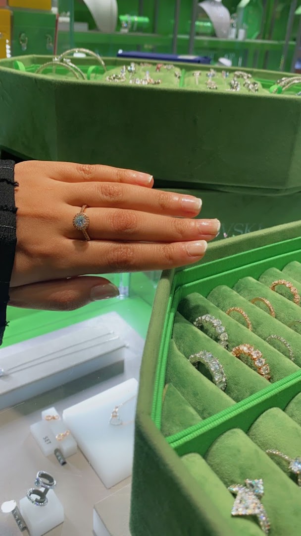 A hand wearing a sparkling ring is shown next to a display of various rings in a green velvet case.