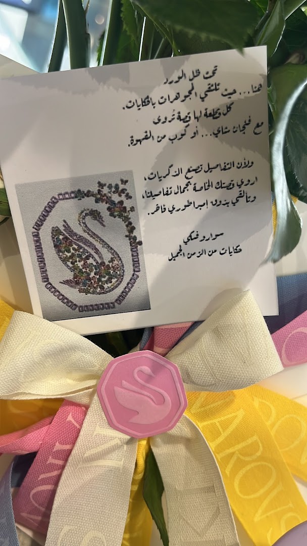 A card with Arabic text and a swan illustration, adorned with colorful ribbons and a Swarovski logo.