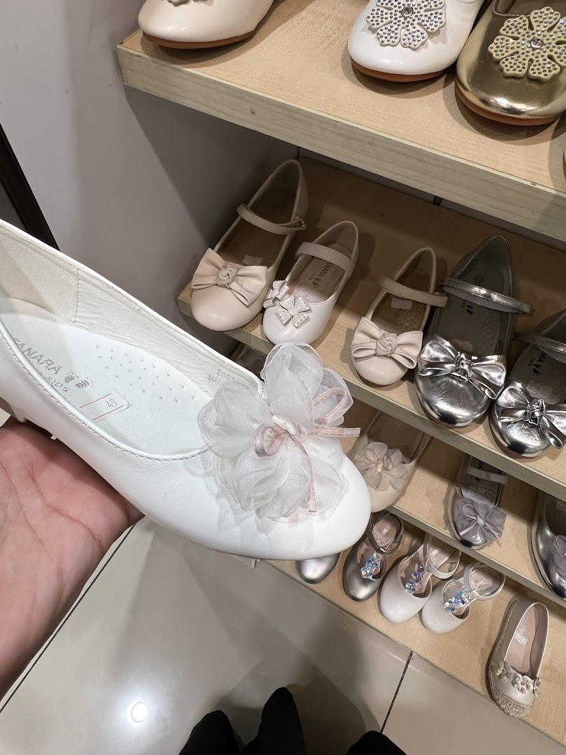 A hand holds a white flat shoe with a large decorative bow, showcasing a collection of children's shoes.