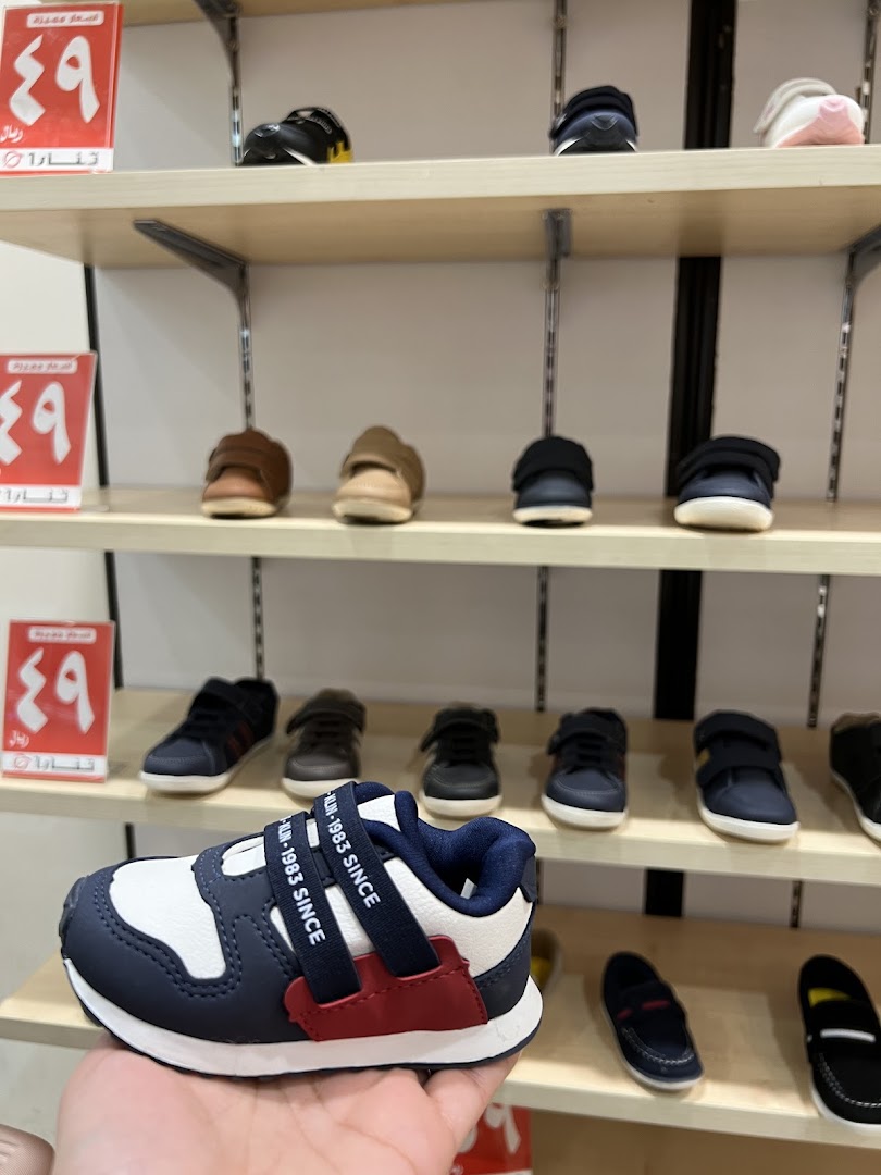 A hand holds a child's sneaker in front of shelves displaying various shoes.