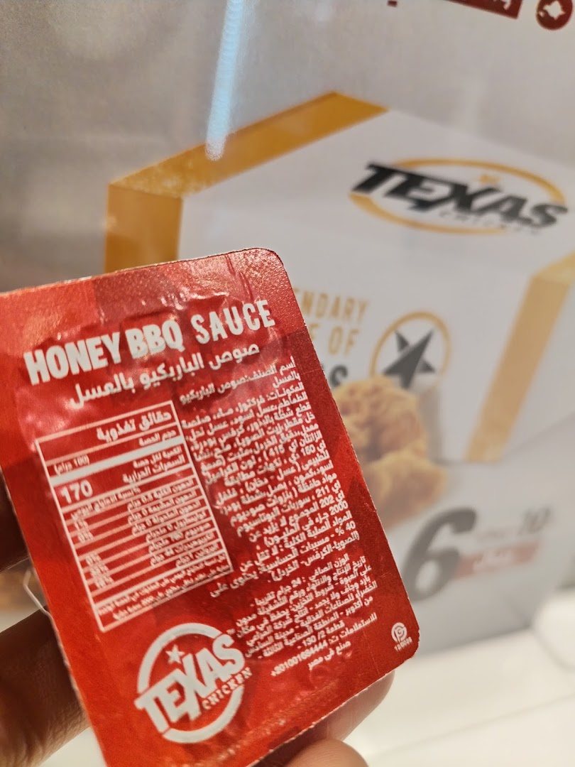 A hand holds a packet of Texas Chicken Honey BBQ Sauce with nutritional information.