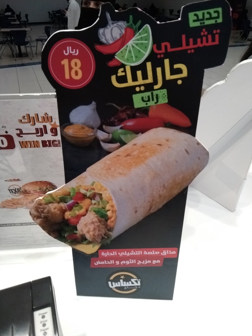 A promotional stand for Texas Chicken featuring a large wrap and advertising a chili garlic wrap for 18 Riyals.