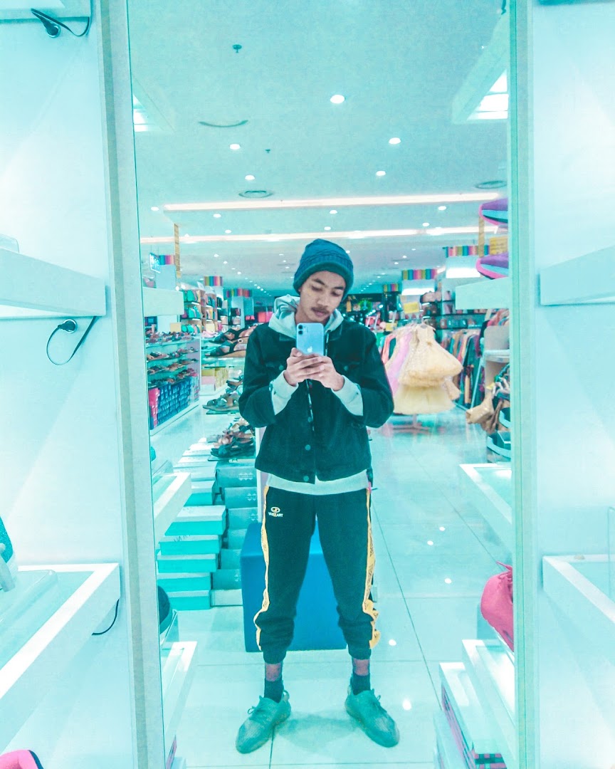 A young person in a beanie and casual wear takes a mirror selfie inside a brightly lit store.