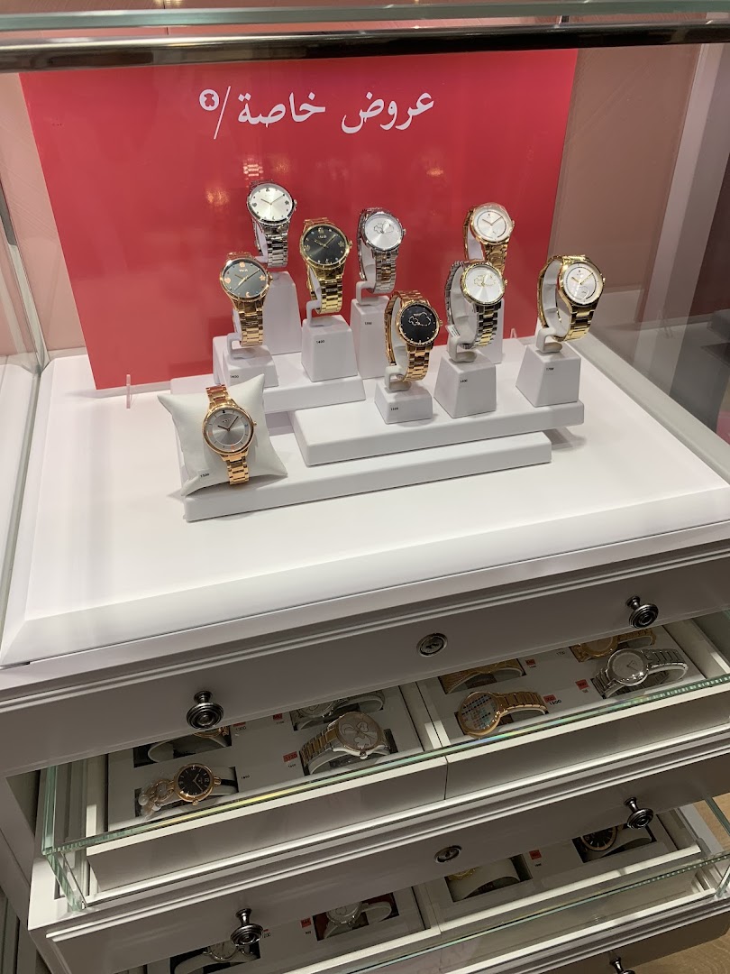 A display of various watches in a shop window, with a red background featuring Arabic text.
