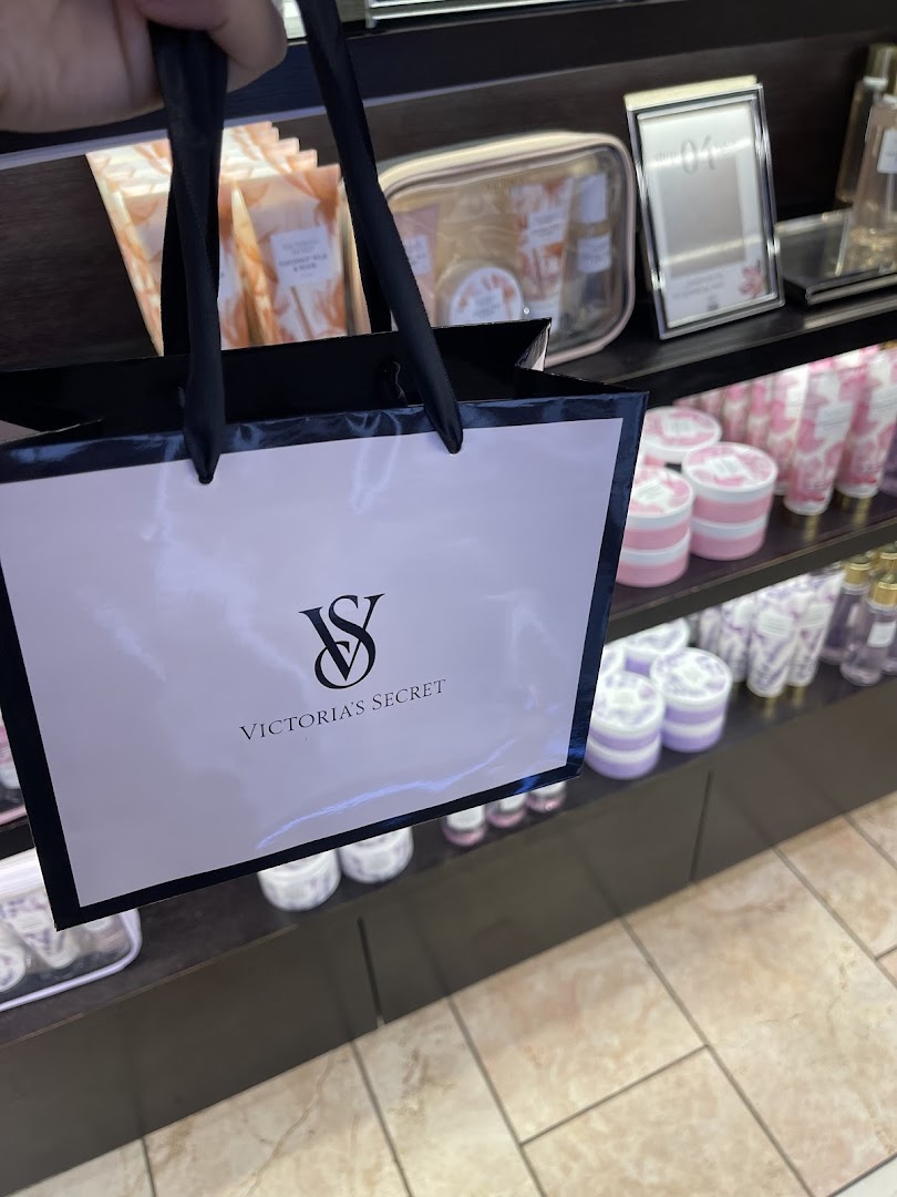 A hand holds a Victoria's Secret shopping bag in front of shelves stocked with beauty products.