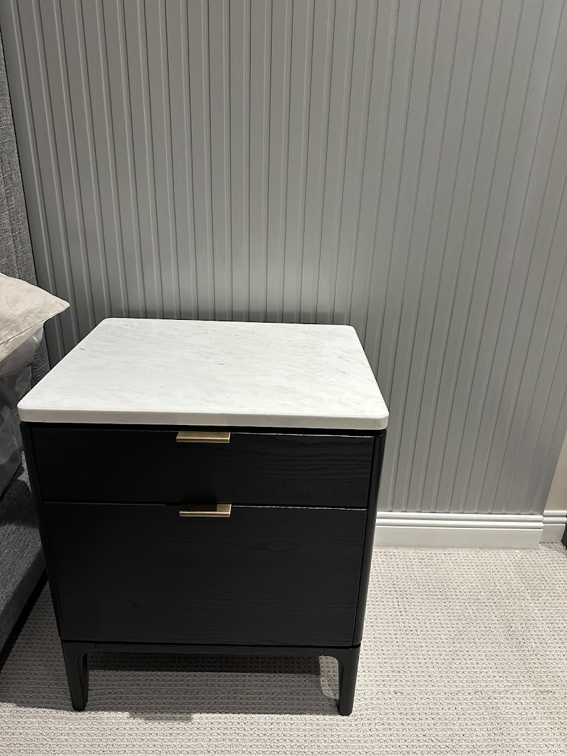 A black nightstand with a white marble top and brass handles sits against a grey paneled wall.