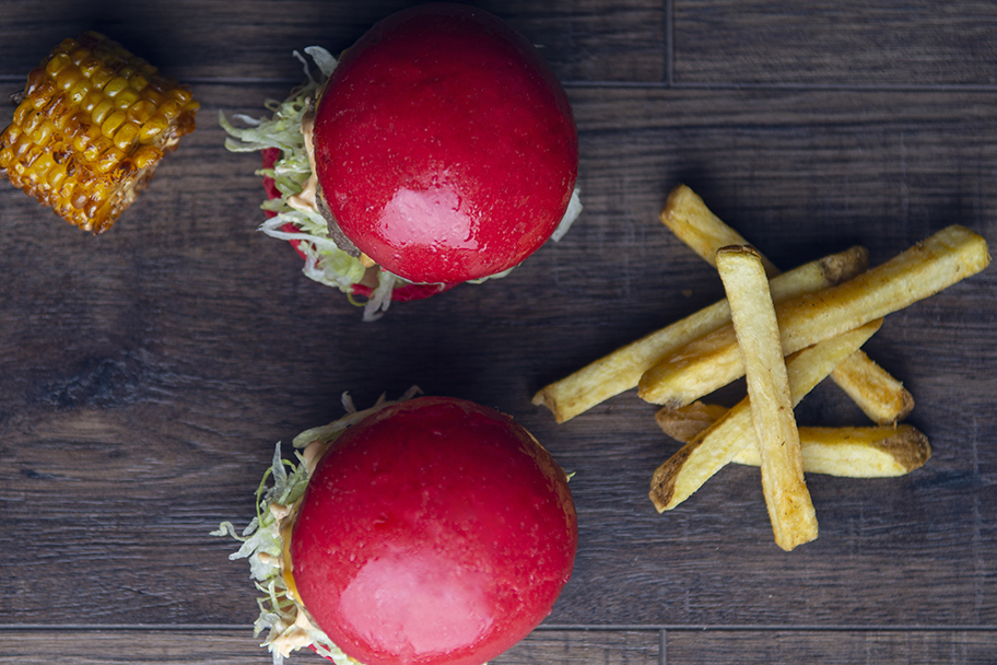 Two red-bun burgers with fries and corn on a wooden table.