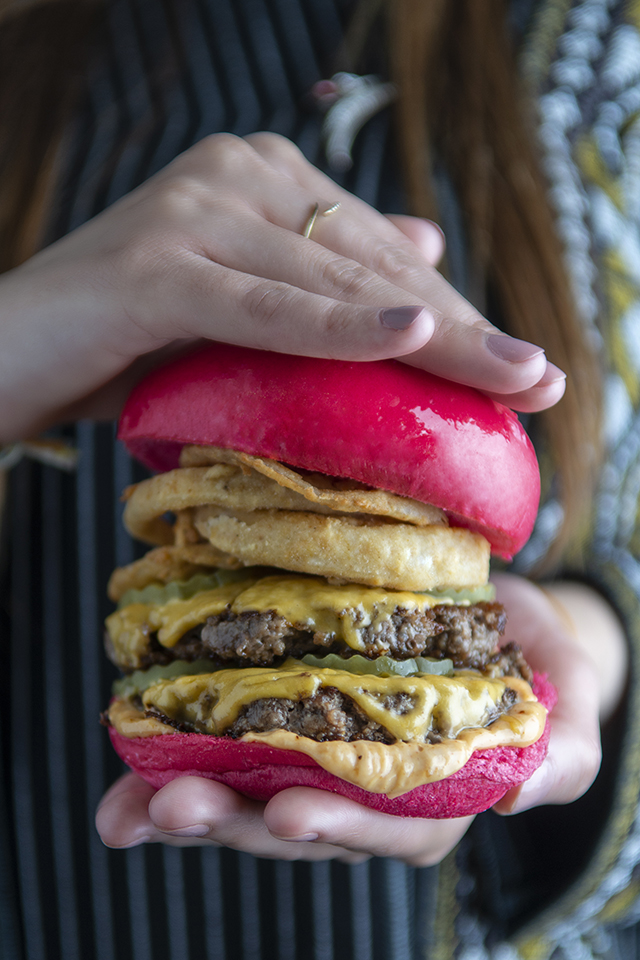 A person holds a large, double-patty burger with pink buns and onion rings.