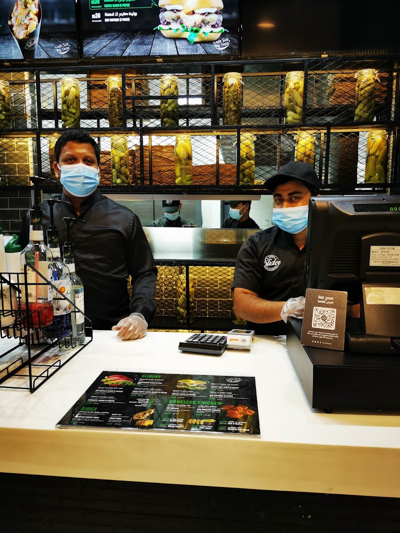 Two staff members wearing masks stand behind a counter with a menu and jars of pickles.
