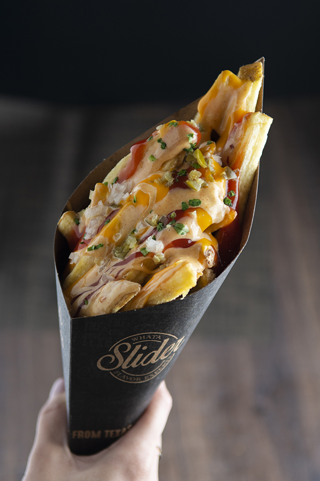 A hand holds a cone of loaded fries with cheese, sauce, and toppings.
