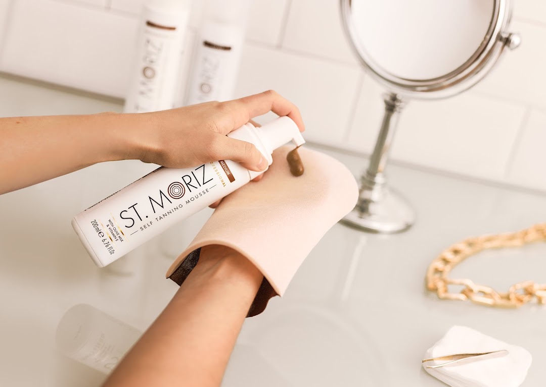 A person applies self-tanning mousse to a tanning mitt with a mirror in the background.