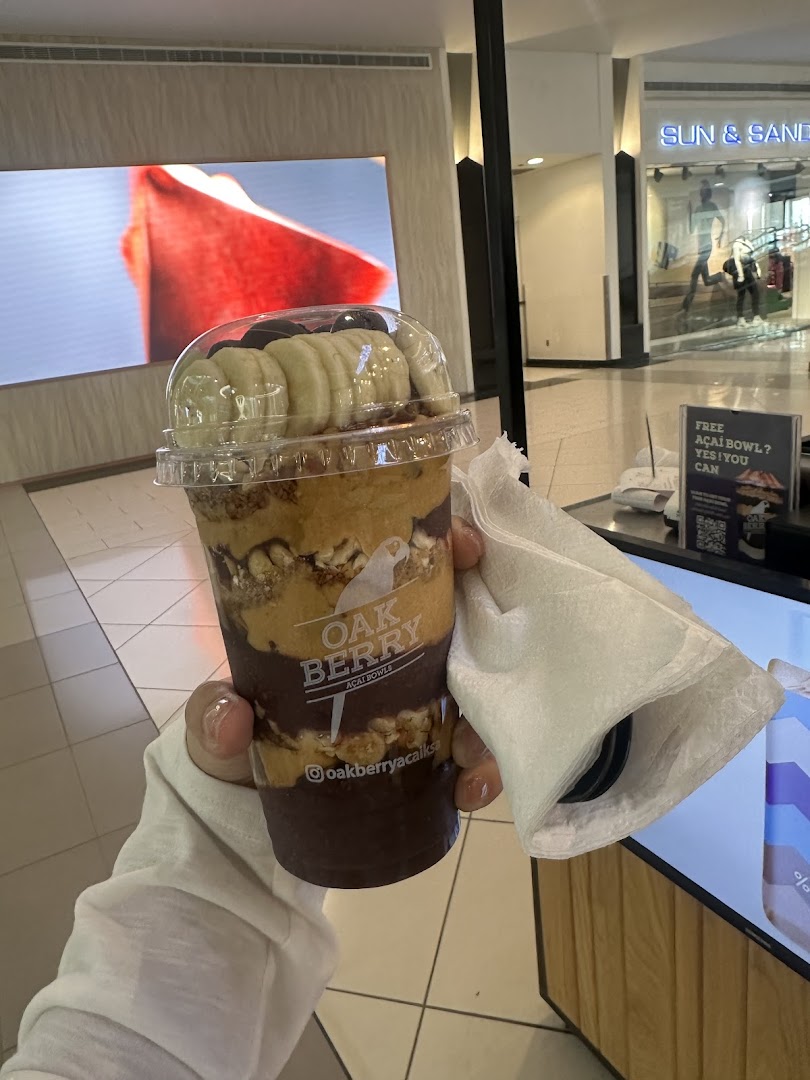 A person holds a large acai bowl with bananas and granola, with a mall background.